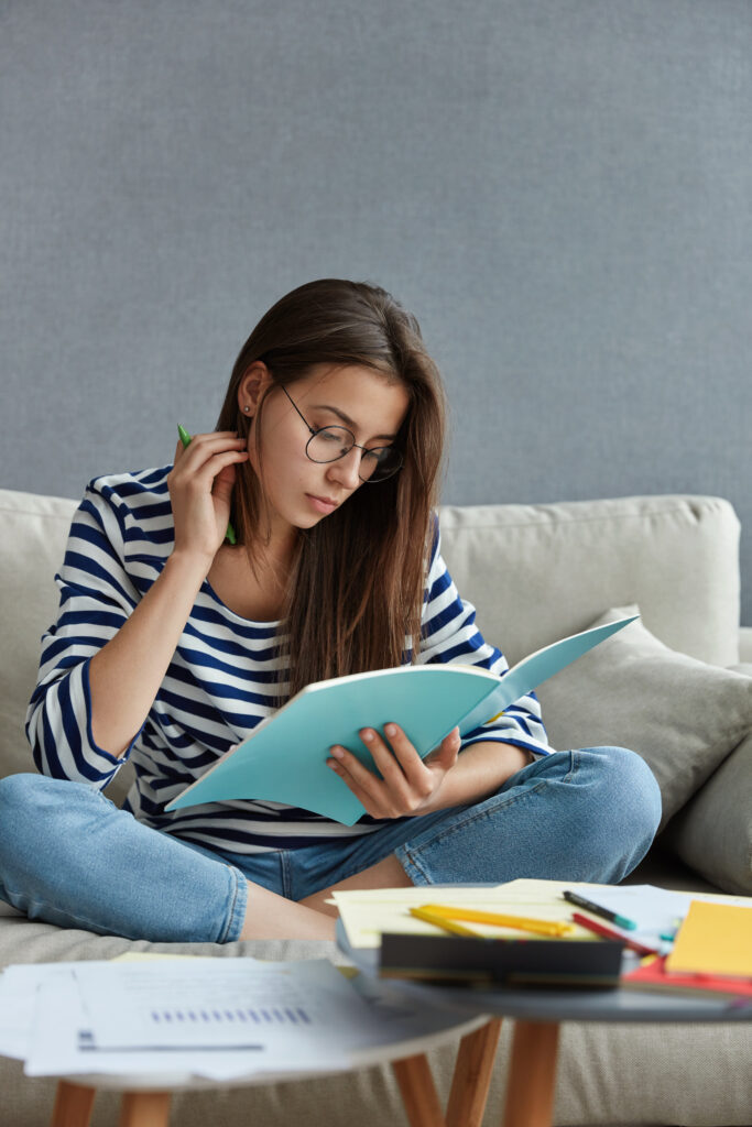 pretty student with dark hair focused in textbook, prepares for writing course paper or upcoming exam, sits crossed legs on comfortable sofa against modern apartment interior. studying concept pretty student with dark hair focused in textbook, prepares for writing course paper or upcoming exam, sits crossed legs on comfortable sofa against modern apartment interior. studying concept